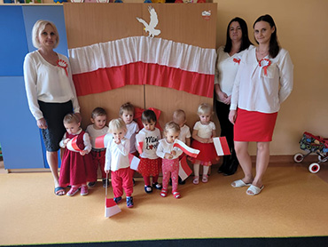Dzieci stoją razem z Paniami ubrane w stroje biało – czerwone i trzymają biało – czerwone flagi