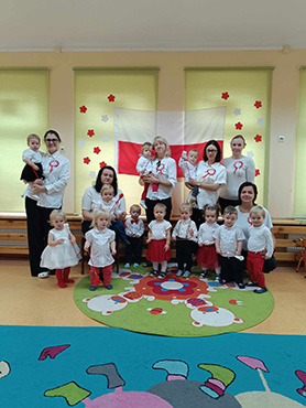 Dzieci stoją razem z Paniami ubrane w stroje biało – czerwone i trzymają białe lub czerwone kwiatki