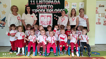 Dzieci stoją razem z Paniami, w pierwszym rzędzie dzieci siedzą. Wszystkie dzieci ubrane w stroje biało – czerwone i trzymają flagi