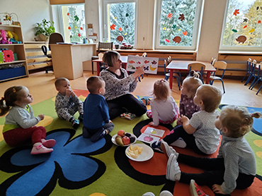 Zdjęcie przedstawia grupę dzieci i opiekunkę, która czyta opowiadanie