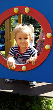 Zdjęcie przedstawia uśmiechniętą dziewczynkę, która zagląda przez kółeczko na placu zabaw.