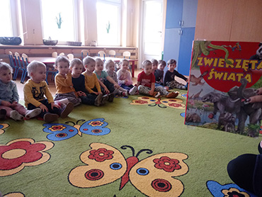 Dzieci siedzą w grupie, pani trzyma książkę z napisem „Zwierzęta Świata”