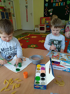 Dwóch chłopców siedzi przy stoliku i maluje farbami makaron