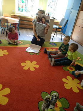 Pani pokazuje zdjęcie dzieciom; dzieci siedzą w kle na dywanie