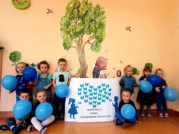 Zdjęcie grupowe. Dzieci trzymają w rękach niebieskie balony. Dwoje dzieci trzyma wykonaną pracę plastyczną.