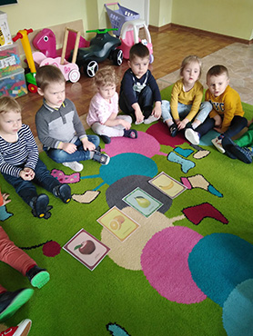 Dzieci siedzą na dywanie a przed nimi rozłożone są ilustracje znanych owoców
