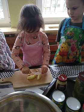 Dziewczynka kroi na deseczce obrane jabłko, a obok niej stoi chłopczyk