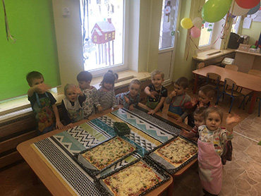 dzieci stojące wokół stolika, na którym leżą trzy blachy z gotowymi do pieczenia pizzami
